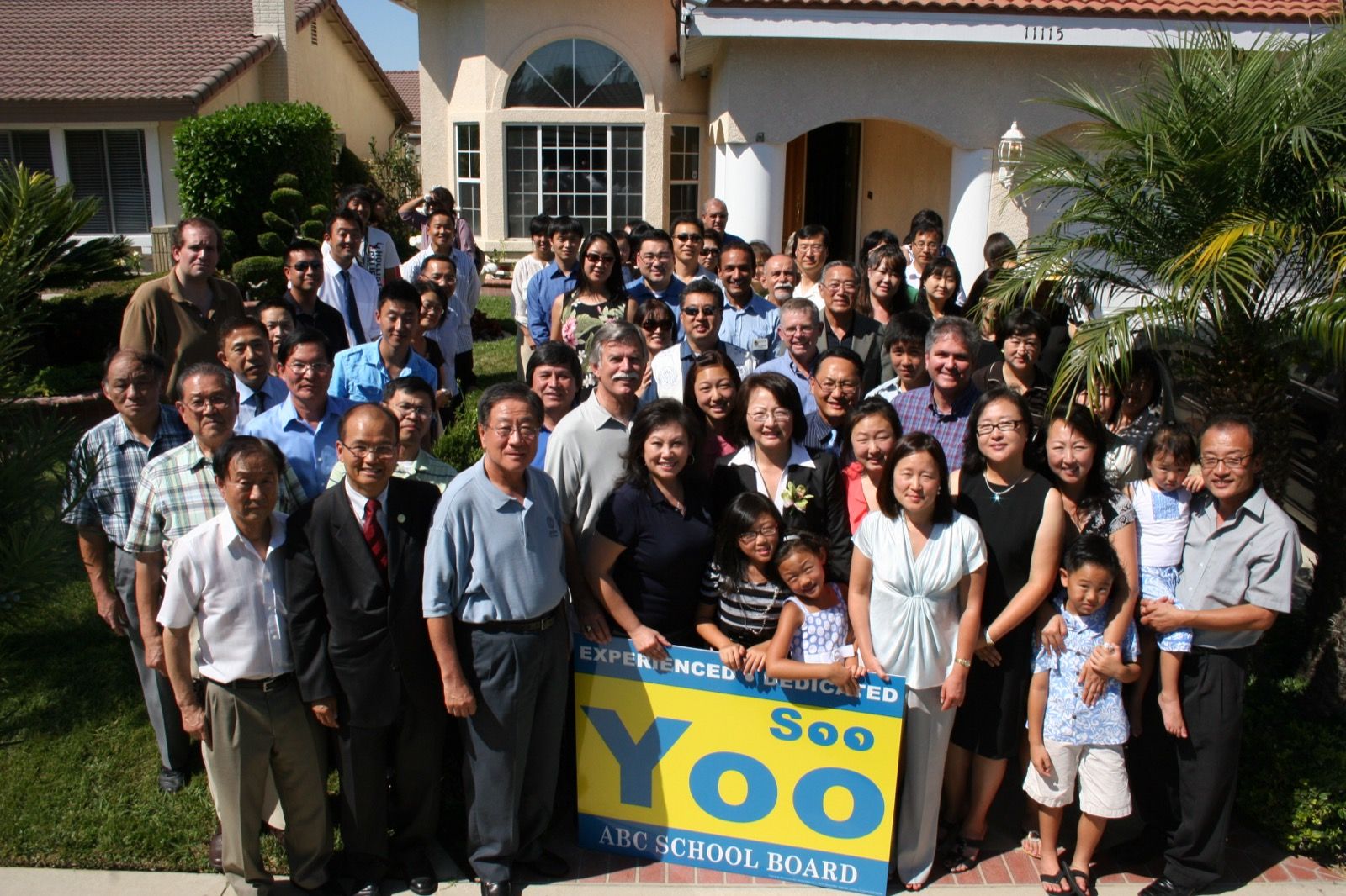 Soo Yoo with a large group of supporters gathered outside a home behind a yellow and blue “Soo Yoo, ABC School Board” campaign sign